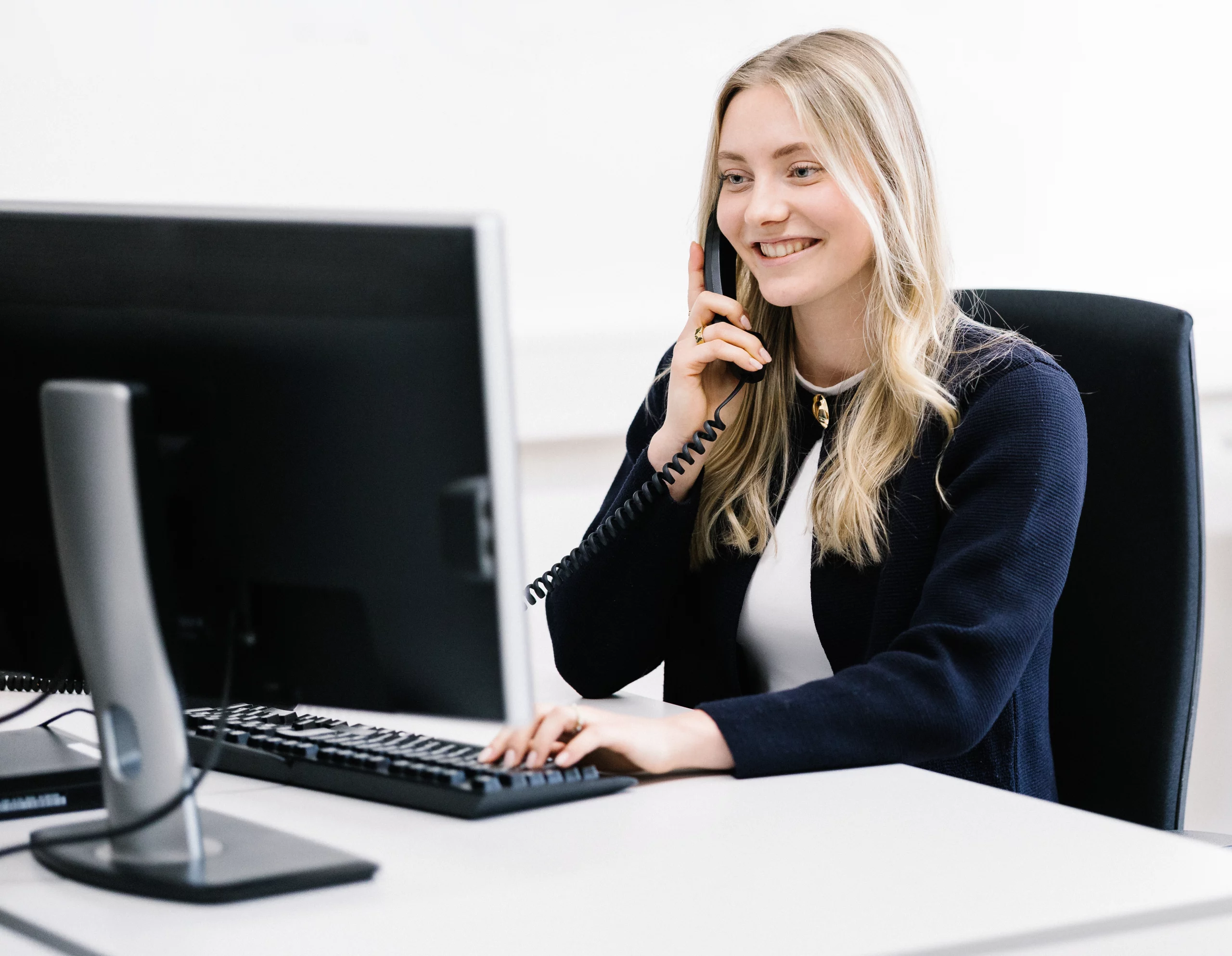 Lächelnde Mitarbeiterin sitzt am Computer und telefoniert an ihrem Arbeitsplatz.