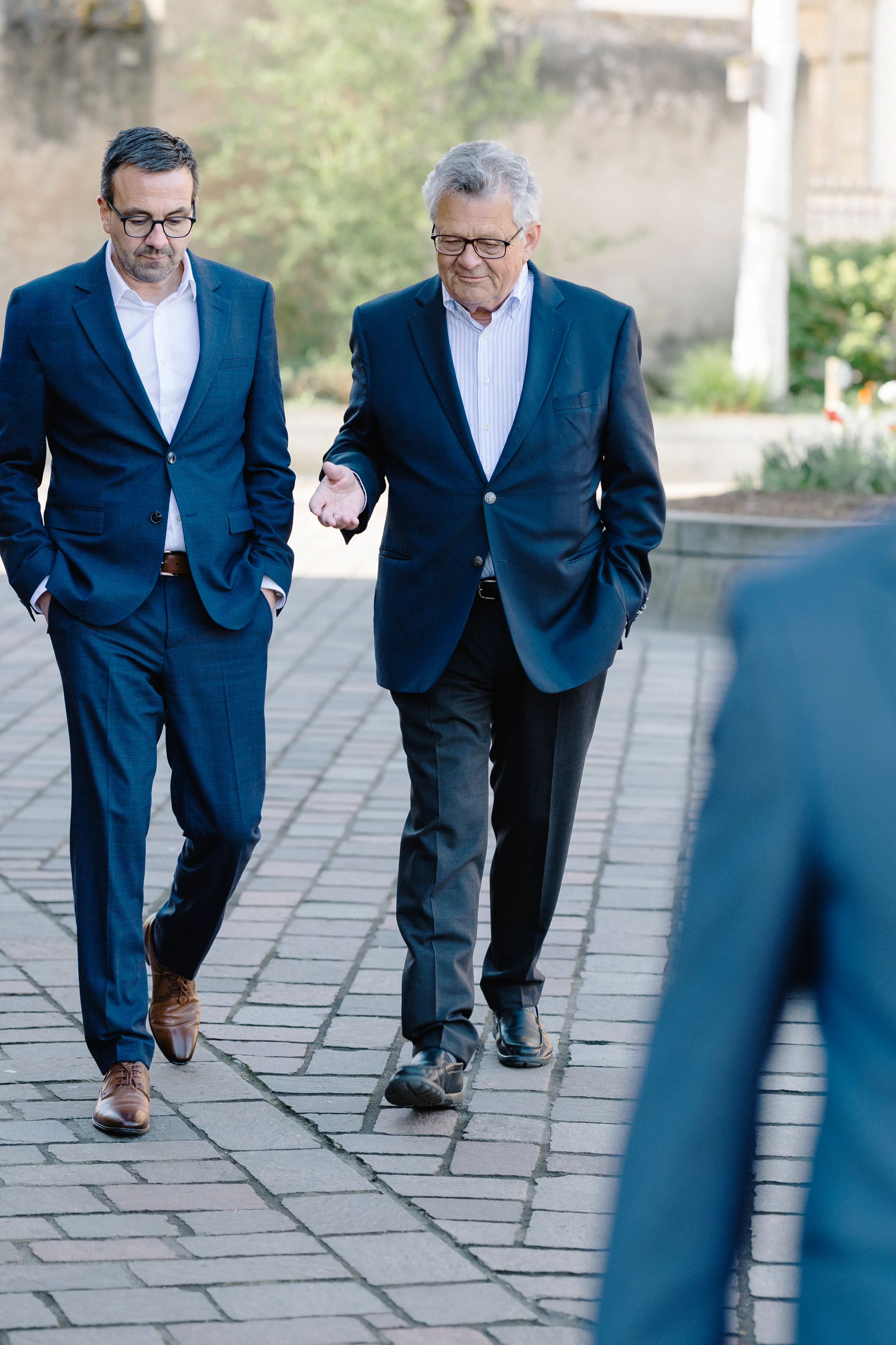 Zwei Männer gehen gemeinsam im Gespräch über einen gepflasterten Weg.